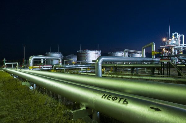 Pipelines on the background of a processing plant. On the tube, it is written in Russian the word "oil." At night, the different light sources are concentrated in different parts of the plant, creating different shades of color refinery.